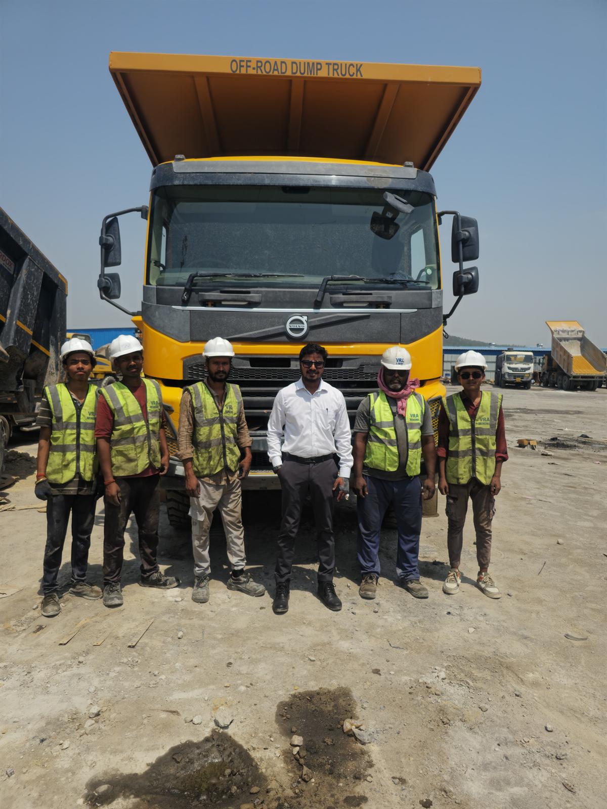 VRA on-site team wearing safety vests and white hard hats standing in front of a yellow Volvo dump truck